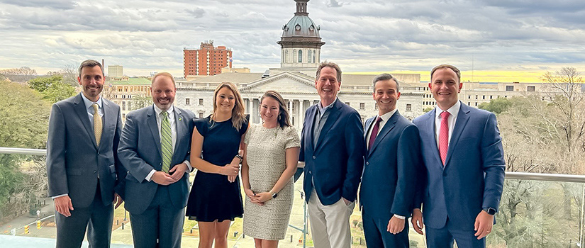 Nikki Crabtree Huber poses in the center of seven people with the South Carolina Statehouse in the background.