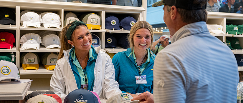 Two students working in merchandising at the Masters hand a customer a hat.