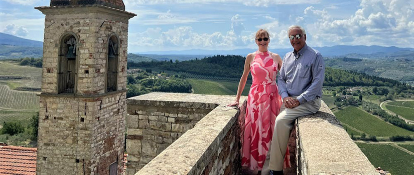 Harris and Patricia Pastides pose for a atop a wall with a bell tower to the left and the landscape of Italy in the background.