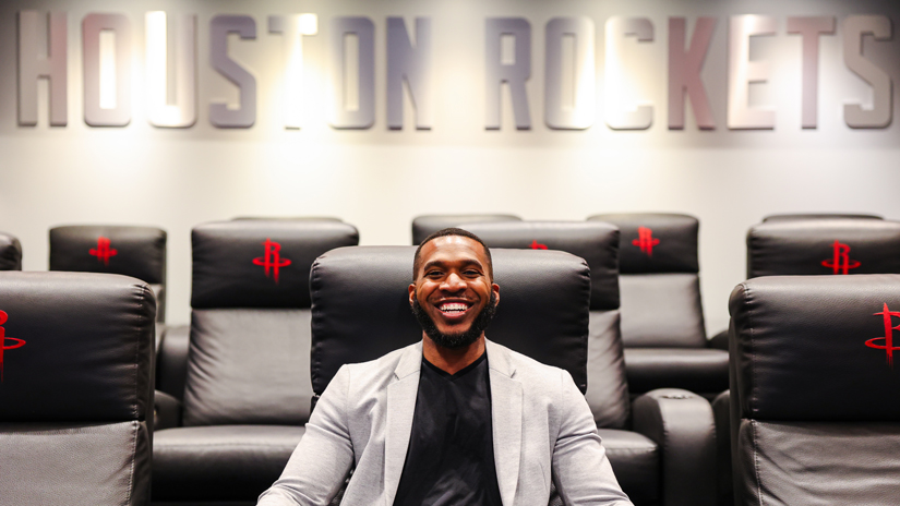 Duane Scott poses in a chair with Houston Rockets displayed on a wall in the background