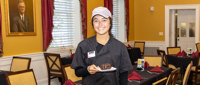 Hospitality management student Josie Anaya smiles while holding a dessert plate inside USC’s McCutchen House restaurant, where she helps manage operations as part of her coursework.
