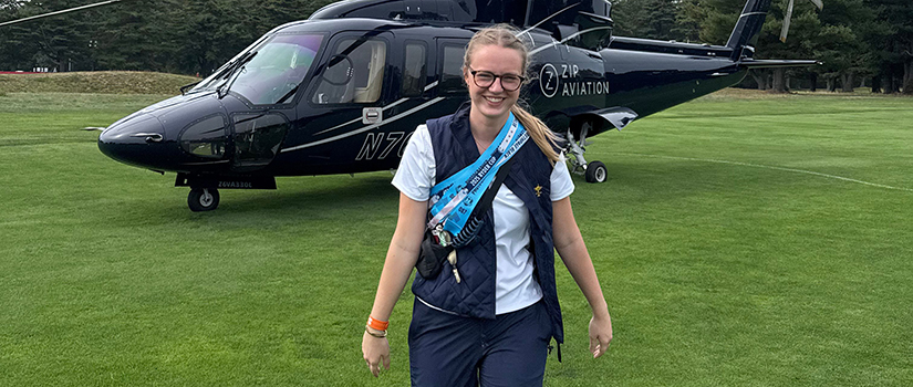 Caitlin Cassidy walks toward the camera smiling after stepping away from a black Ryder Cup transport helicopter behind her.