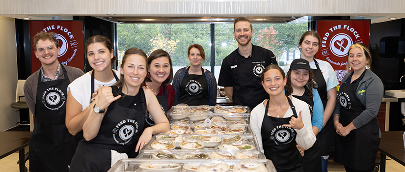 Group of USC students and faculty volunteers smiling behind tables stacked with packaged meals for the Feed the Flock food sustainability initiative.