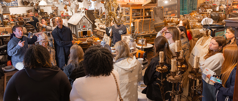 A group of USC retailing students listens as two antique dealers speak inside a packed High Point Market showroom filled with vintage furniture, lighting fixtures and décor.