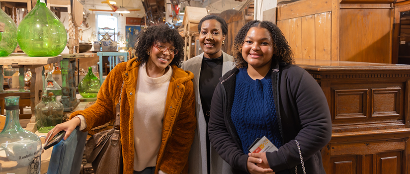 Two USC retailing students and staff member Lena Pinkston smile while standing inside an antique shop at the High Point Market, surrounded by green glass bottles, wooden furniture and decorative pieces.