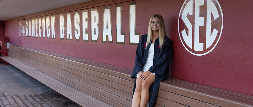 Allison Maloney in a black commencement gown sits along the bench inside the Gamecock Baseball dugout, smiling as she poses beside the “Gamecock Baseball” lettering and large SEC logo on the maroon wall.