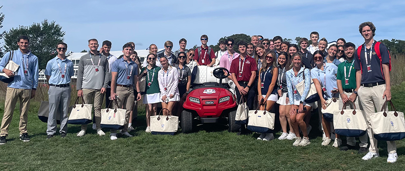 More than 30 HRSM students gather around a red golf utility cart at the 2025 Ryder Cup, holding matching tote bags and wearing event badges.