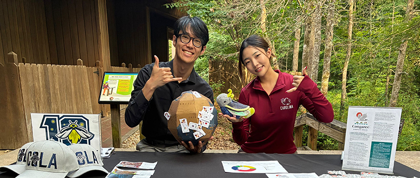 Two University of South Carolina students representing the School of Hospitality and Tourism Management sit behind an information table at Congaree National Park, displaying materials and educational items related to a firefly tourism research project.