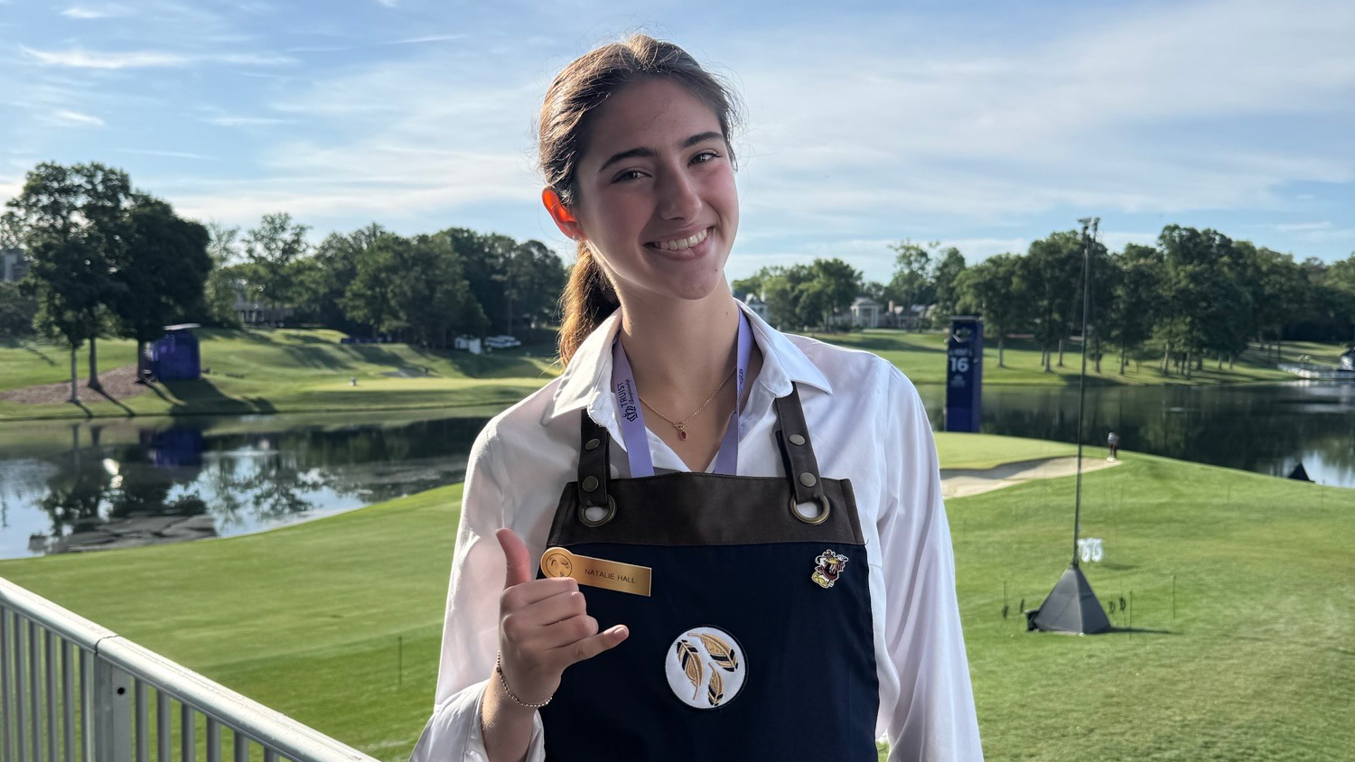 Natalie Hall, a University of South Carolina hospitality management student, smiles while wearing a navy apron and white shirt on a balcony overlooking a golf course and lake.