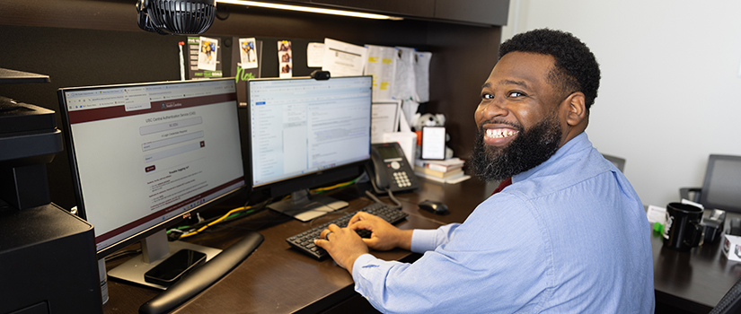 HRSM academic advisor Martiez Moore smiles while working at his dual-monitor workstation, where he uses artificial intelligence tools and advising technology to streamline administrative tasks and create more time for meaningful conversations with students.