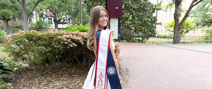 University of South Carolina hospitality management graduate Kaila Regan smiles on campus wearing her graduation stole recognizing academic achievement and involvement in the Club Management Association of America.