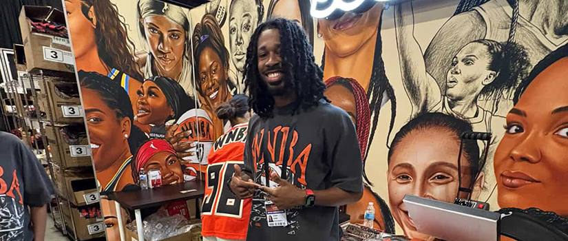 Chadd Rowe stands inside a merchandise booth wearing a black WNBA T-shirt, with colorful jerseys and apparel displayed nearby. Behind him, a large mural features illustrated portraits of women's basketball players in vibrant colors.