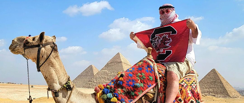 Research professor Rich Harrill holds a University of South Carolina Fighting Gamecocks flag while sitting atop a camel with the pyramids of Egypt in the background.