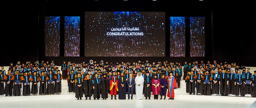 Graduates from HBKU in Doha, Qatar, pose on a stage with the worlds Congratulations on a videoboard in the background.