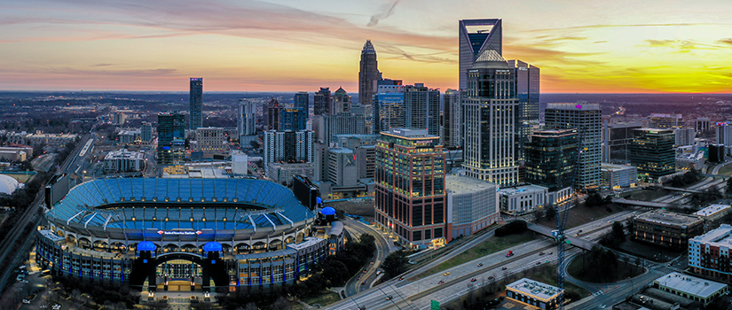 Aerial panoramic view of Uptown Charlotte at sunset featuring Bank of America Stadium in the foreground, the city skyline illuminated against a colorful evening sky, surrounding highways and modern office buildings visible throughout the urban landscape.