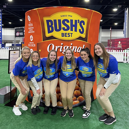 USC beach volleyball player Kaeli Crews poses for a photo with five other students in front of a large Bush's Baked Beans sign.