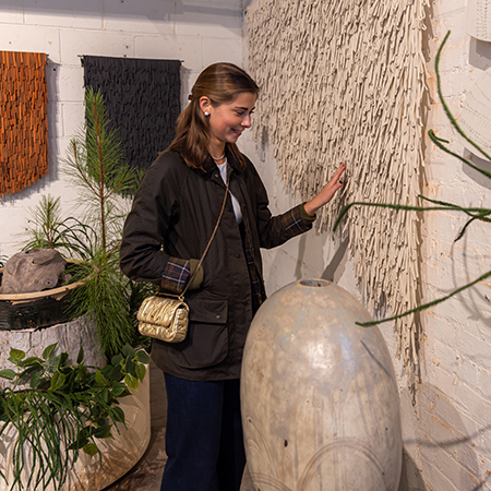 A USC retailing student examines a textured wall art installation inside a High Point Market gallery featuring natural materials, plants and sculptural pieces.