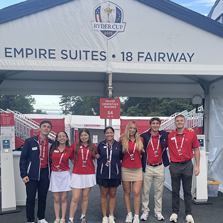 A group of HRSM students in red and navy uniforms pose together outside the Empire Suites entrance at the 18th Fairway during the 2025 Ryder Cup.