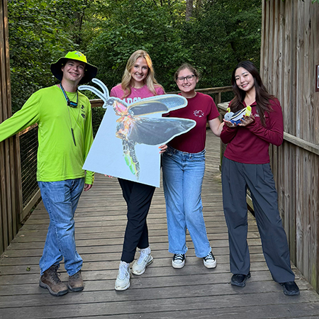 A group of University of South Carolina students and partners stand on a wooden boardwalk at Congaree National Park holding a large firefly display and educational materials during a tourism research initiative.