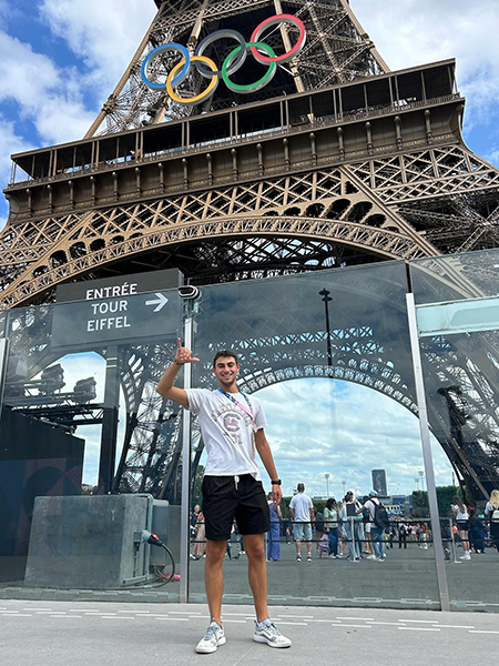 Trevor Lazarus stands in front of the Eiffel Tower decorated with Olympic rings.