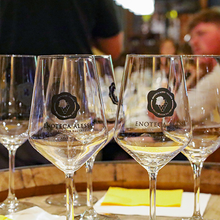 A collection of wine glasses with the text "Enoteca Alessi Delicatessen Dal 1952, Firenze" in the foreground and patrons sitting at a table in a blurred background.