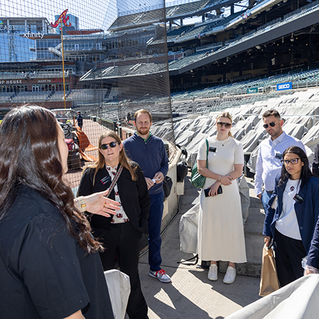 A group of students and professionals stands inside a baseball stadium while listening to a guided tour. The group gathers beside the seating area near the field as a speaker explains the venue, providing a behind-the-scenes learning and networking experience in a sport industry setting.