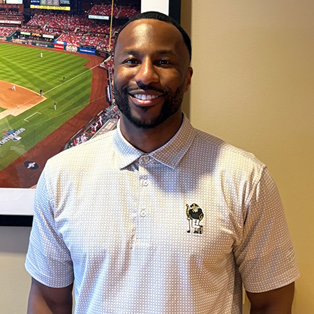 A sport industry professional smiles for a portrait while standing indoors in front of a framed photograph of a baseball stadium.