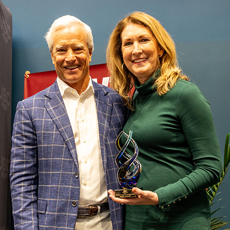 Two people pose together at an awards ceremony, smiling toward the camera. One person holds a glass award with a blue spiral design while standing beside a presenter in a blue checkered blazer. A branded event backdrop and stage elements are visible in the background.