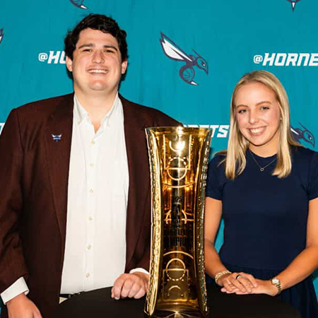 Two students pose beside a championship trophy at a professional sports venue. Standing in front of a team-branded backdrop, they smile toward the camera while showcasing the trophy, highlighting hands-on industry access and experiential learning opportunities in sport and entertainment management.