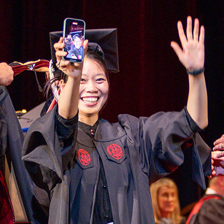 A smiling graduate in a black cap and gown waves to the audience while holding up a smartphone to take a selfie during a commencement ceremony. The phone screen shows a video call or selfie view as the graduate celebrates crossing the stage. Other graduates and ceremony participants are visible in the background.