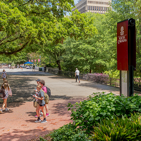 Students walk along a shaded campus pathway near a University of South Carolina wayfinding sign.