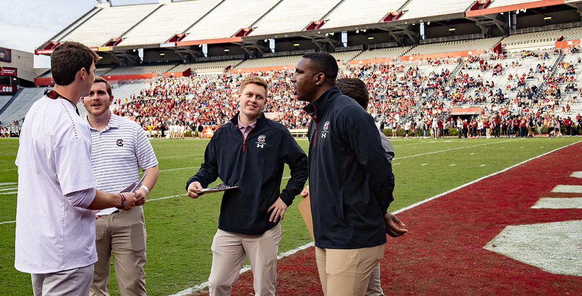 A group of five students discuss their work assignments on the field at Williams-Brice Stadium prior to the Garnet and Black Spring Game in April 2022.