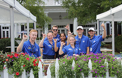 HRTM students working the PGA tournament at Charlotte's Quail Hollow Club pose signalling the "spurs up!" hand sign while standing in front of the clubhouse.
