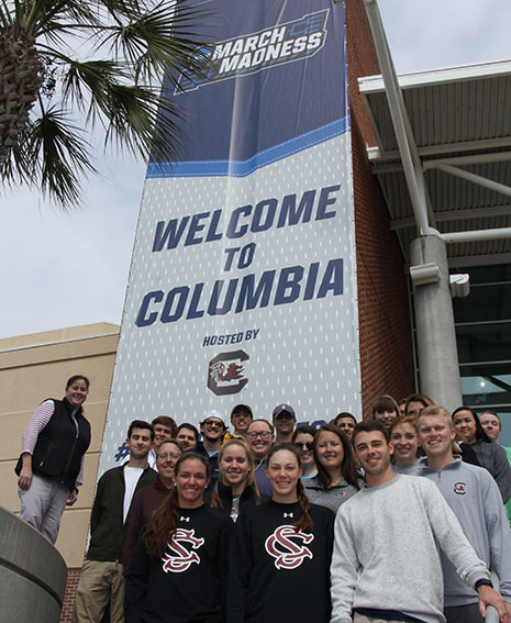Students in Haylee Mercado's sport and entertaiment management class pose in front of the March Madness banner outside Colonial Life Arena. Students were able to go behind the scenes to learn about event operations at the tournament.