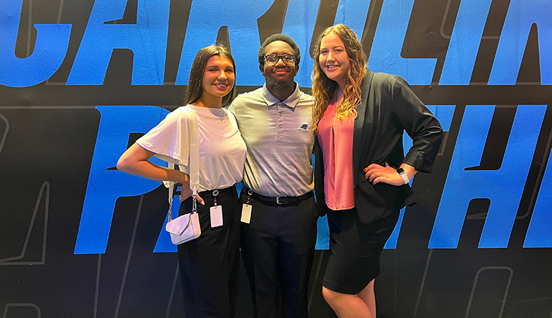 Three Tepper Scholars pose for a photo with the Carolina Panthers logo in the background.