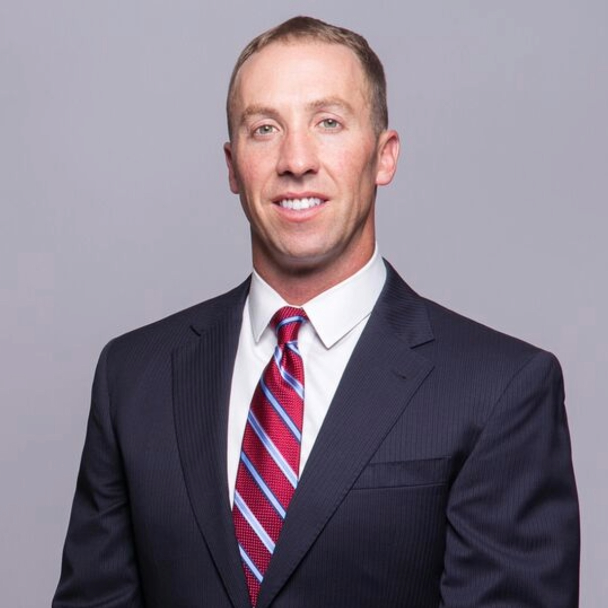 Micah Kurtz dressed in a dark business suit and red striped tie poses for a professional headshot against a gray studio background.