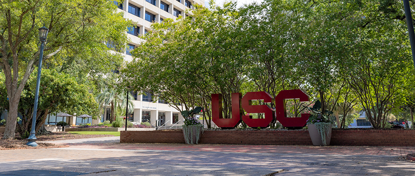 Large red USC letters displayed in a landscaped plaza outside a campus building, with mature trees providing shade and a pedestrian walkway leading through the foreground.