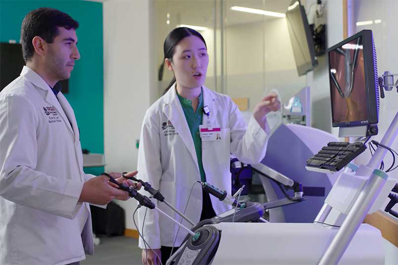 Female instructor watches a monitor as med student manipulates robotic handles.