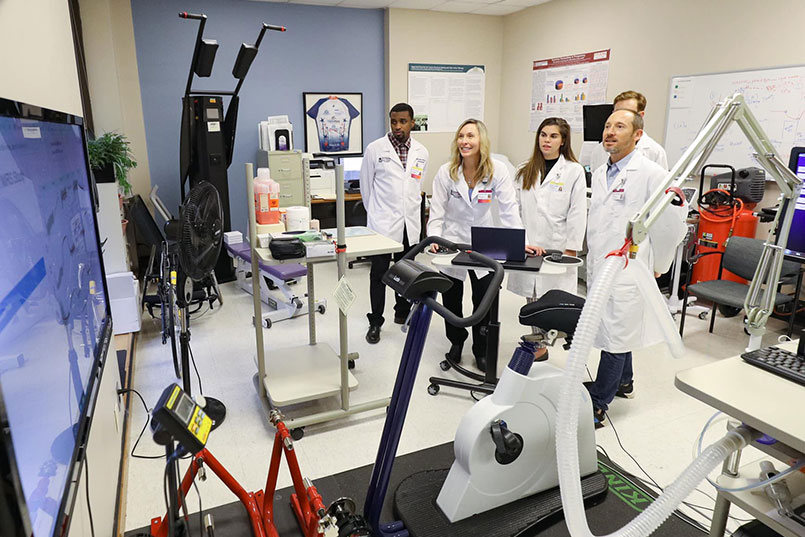 Medical students gather around a professor in a lab full of equipment who demonstrates how display data on a large monitor.