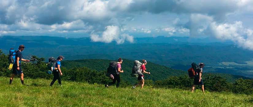 Hikers walk along a grassy ridge with mountains in the background.