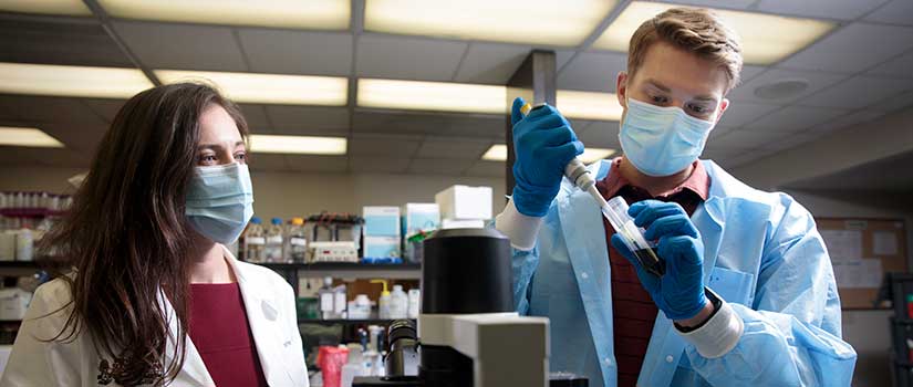 Student in mask and blue labcoat fills pipette while mentor observes.