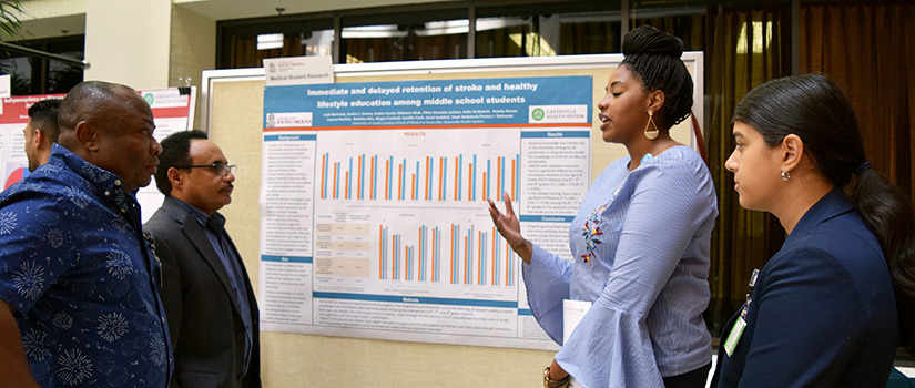 Two female med students explain their research poster to onlookers.