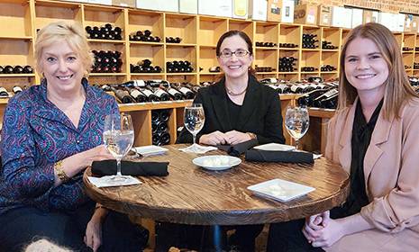 Karen Brosius, Christina Ahmadjian, Folks Center Sonoco Visiting Fellow, and Madison Buckles, Folks Center Program Coordinator, sitting at a table; bottles of wine are stored on shelves in the background