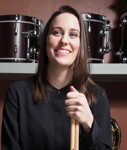 Woman with medium length dark hari, wearing black and holding drum sticks. Behind her are drums on shelves.