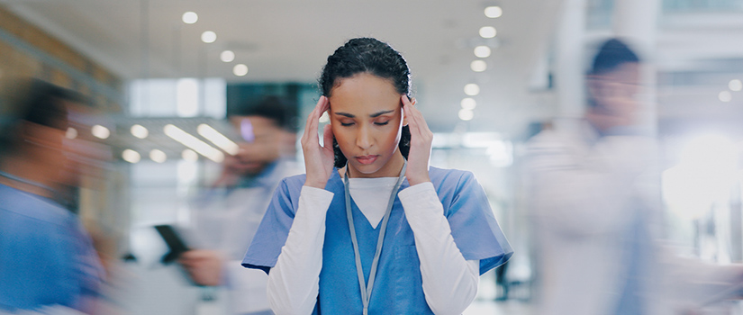 nurse in scrubs holds her temples while blurred hospital activity moves around her, suggesting stress in a busy workplace