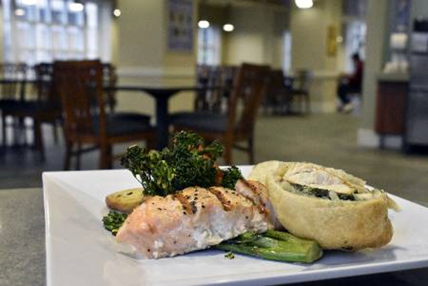 A a plate of baked salmon, chicken cordon bleu and broccolini on a table with other tables in the background.