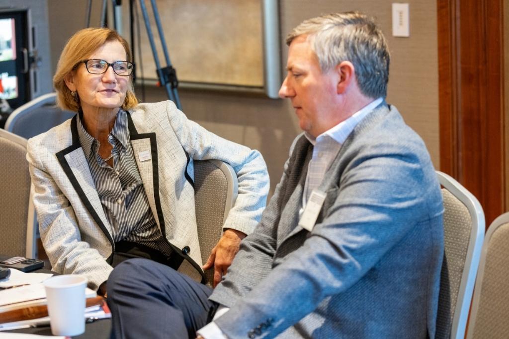 A man and woman in suits speak at a table. 