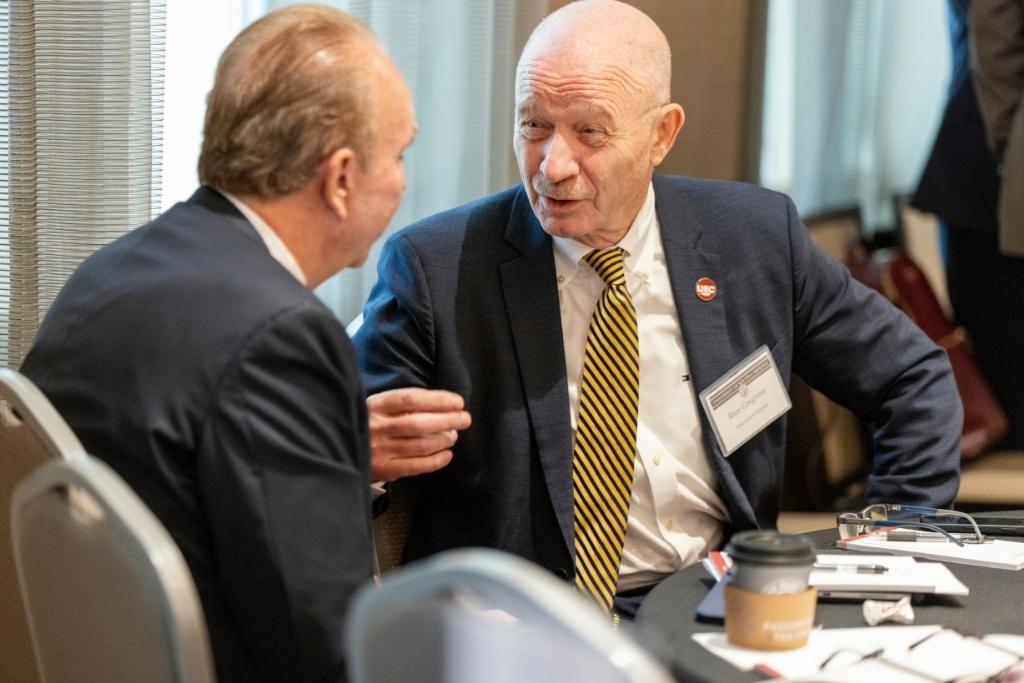Two men in suits speak together at a table. 