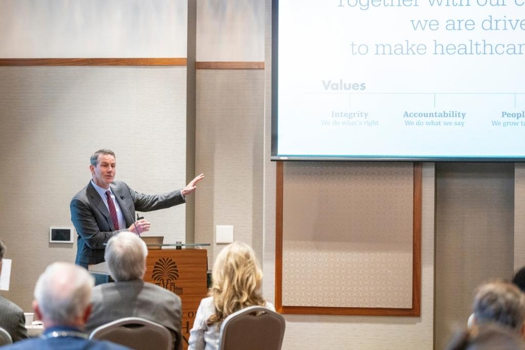 A man in a suit gives a presentation, pointing at the projector screen as a conference room full of people listen. 