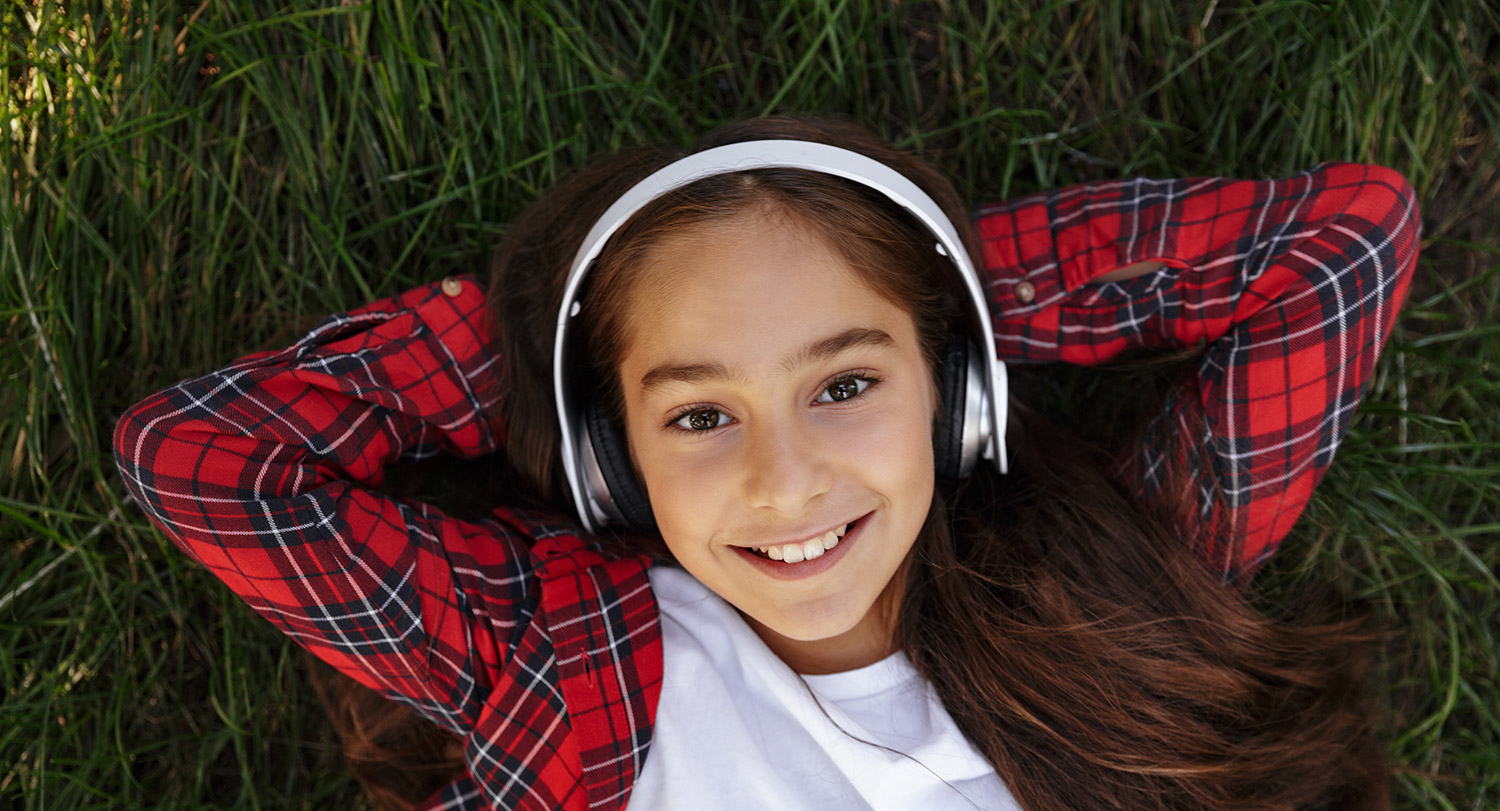Adolescent girl wearing headphones, smiling and relaxing on grass field.
